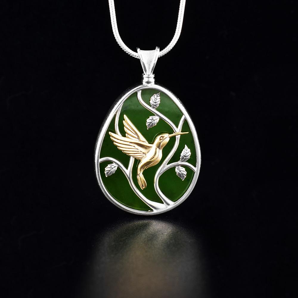 This jade pendant necklace has an oval shaped with a depiction of a Hummingbird flying around a tree. The hummingbird is made from 14K gold and the jade and three designs are set in sterling silver.