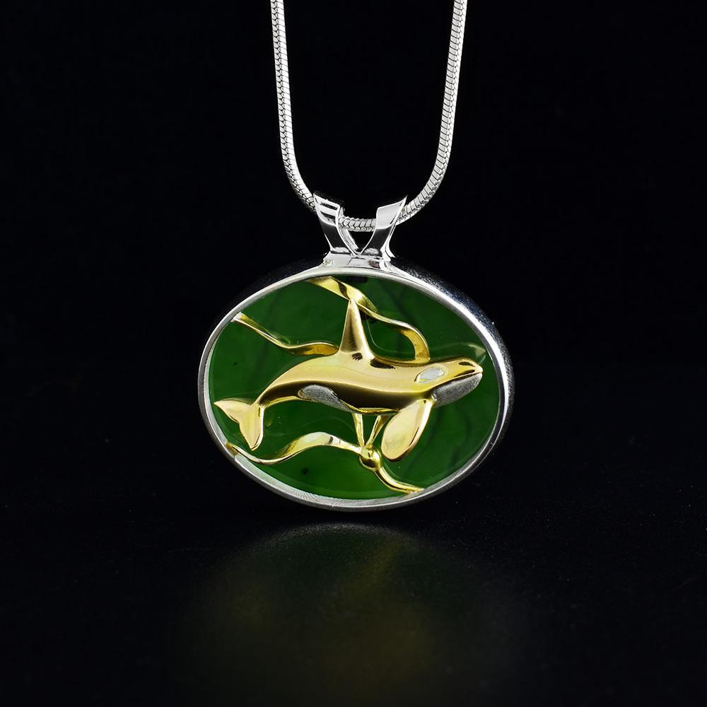 This Orca jade pendant is made from 14K gold, sterling silver, and BC jade. The pendant is oval shaped is made by setting jade into a silver back and has 3D depictions of an orca in kelp made from gold.