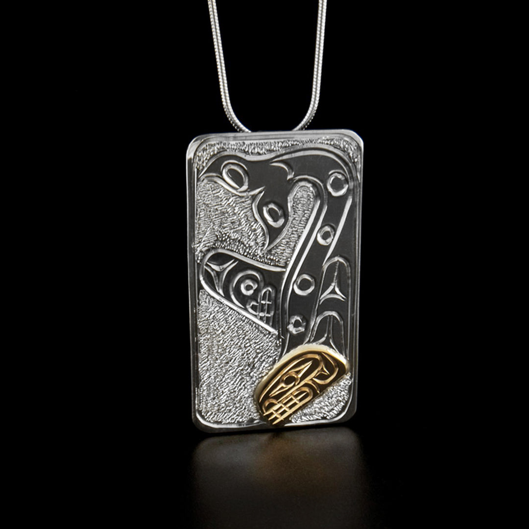 Sterling Silver and 14K Gold Rectangular Pendant by Paddy Seaweed. The design depicts the profile of a full bodied orca upside down. The head of the orca, made from 14K gold, is at the bottom of the pendant looking downward. The body of the orca is above the head with the tail curving upwards at the very top of the pendant. The artist has included intricate designs along the body of the orca. The background has been neatly hand carved to allow for the image of the orca to stand out.