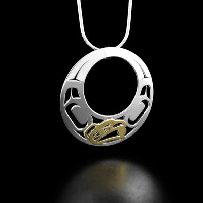 Sterling Silver and 18K Gold Offset Raven Pendant by Grant Pauls. The design depicts the profile of a raven's head made with 18K gold with a small ball in its beak located at the bottom of the pendant. There is a large cutout circle in the middle with intricate designs around it.