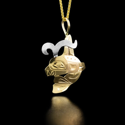 This Gold Orca with Sterling Silver Water Spout Pendant by Fred Myra is in the shape of an orca with the tail right underneath its head. The orca has a wide, open eye looking forward and teeth showing. The artist has handcarved intricate designs along the body to represents the fins and tail. There is a sterling silver water spout coming out of the orca's head.