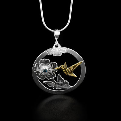 This round hummingbird pendant has a flower in the bottom right with a blue topaz set in the center of it. The hummingbird made from 10k yellow gold to the right of the flower is drinking from it mid-flight.
