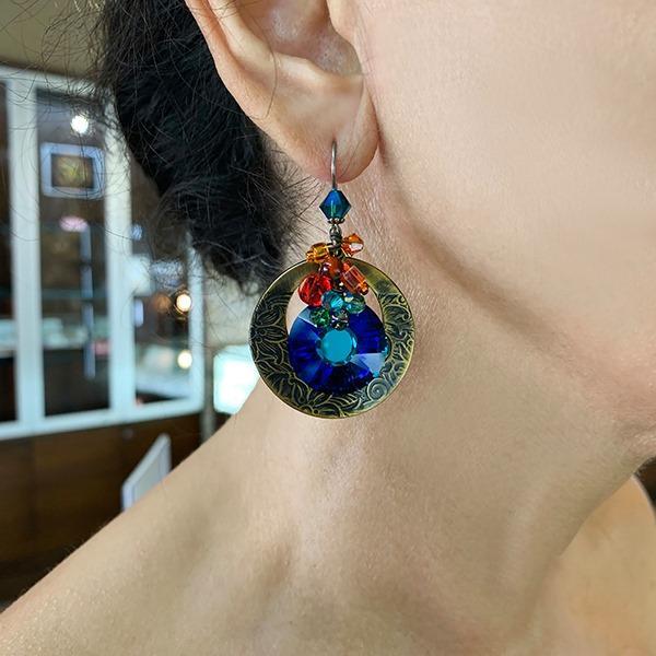 These Swarovski crystal dangle earrings are made from glass, Swarovski crystals and hand worked brass to create this piece. There is a large, circle cutout hangs made from brass with large, dark blue crystals hanging beside them. There are clusters of glass beads and crystals featured on the piece.