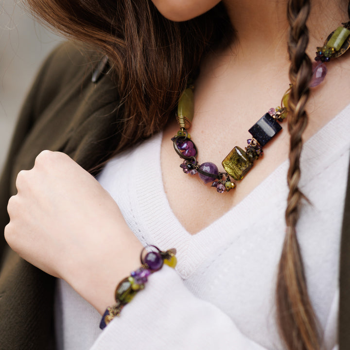 Model with olive-toned skin wearing matching bracelet and necklace. Chunky bracelet made with glass beads, amethyst, jade, cluster crystals, and brass.