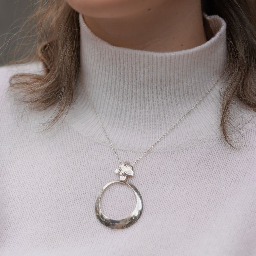 This sterling silver, hollow pendant is a large, tapered hoop attached to a decorative bail. There is a snake skin textured pattern on the surface of the bail.