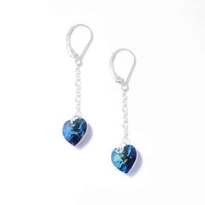 Bermuda Blue Swarovski Crystal Heart Drop Earrings by Debra Nelson. On the end of each earring is a blue heart-shaped Swarovski crystal. The artist has added a sterling silver accent to make the blue colour pop and a long chain leading up to the ear hook.
