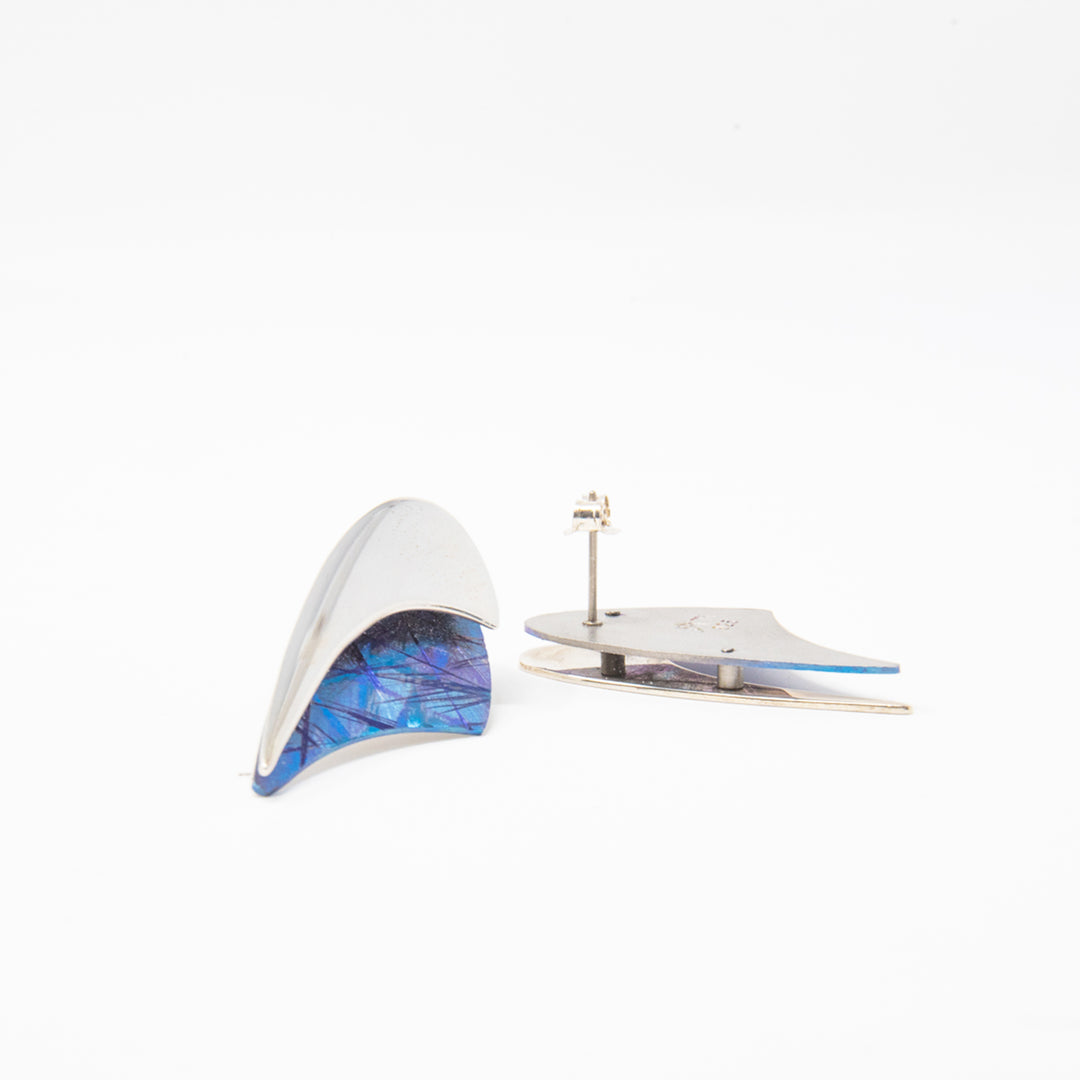 These unique titanium stud earrings are made form sterling silver and anodized titanium. The tops of the earrings are made from a silver band that warps around crescent shaped pieces of titanium. The titanium is blue and has colorful designs on it achieved by anodizing the metal.