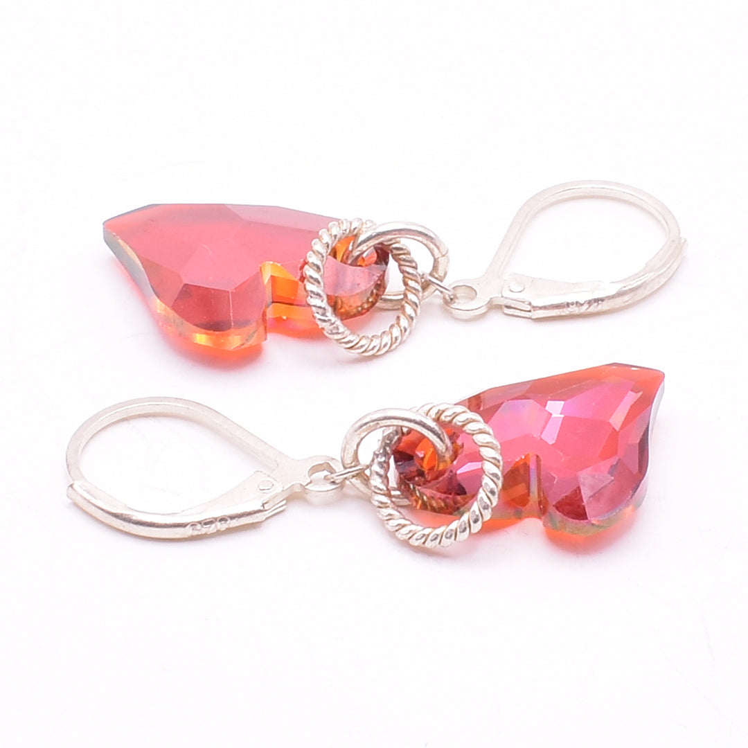 Cinnamon Swarovski Crystal Heart Earrings by Karley Smith. Each earring is in the shape of a lop-sided red heart. The artist has added a sterling silver hoop on top to accent the crystal. The hook is attached to a loop connected to the top of the heart.