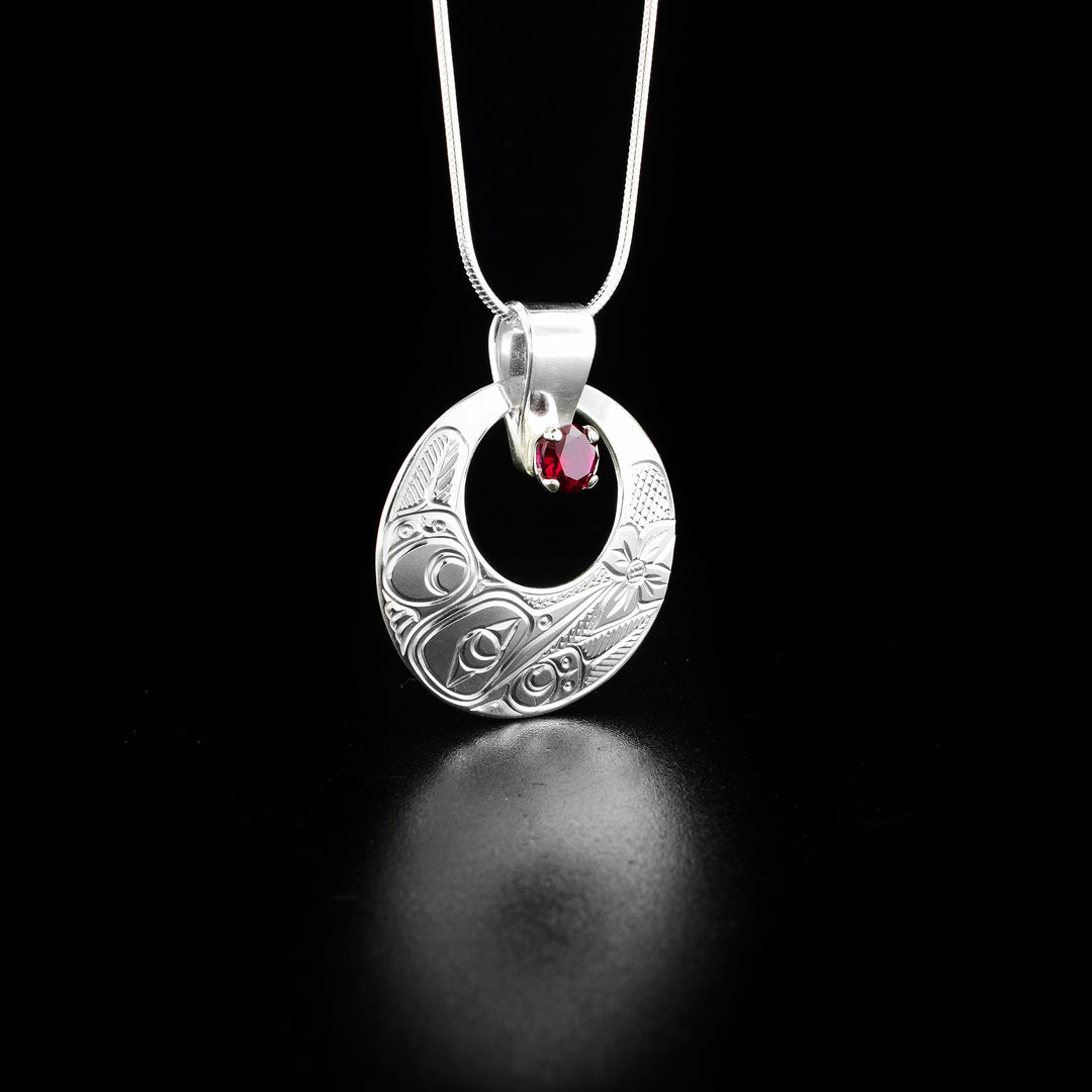 This unique Hummingbird pendant is made from sterling silver and a lab-grown, pink gemstone. The pendant is circle shaped and has a smaller, circle cutout in it. The gemstone is held in the cutout and is attached to the bail. There is a depiction of the Hummingbird carved into the surface of the pendant.