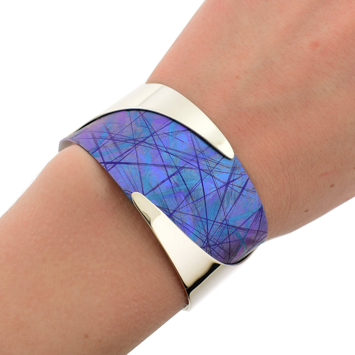 This titanium bracelet is made from sterling silver and anodized titanium and has a single wide band. The titanium is anodized to be blue and has colorful designs, there are wavy, sterling silver highlights running along the bracelet.