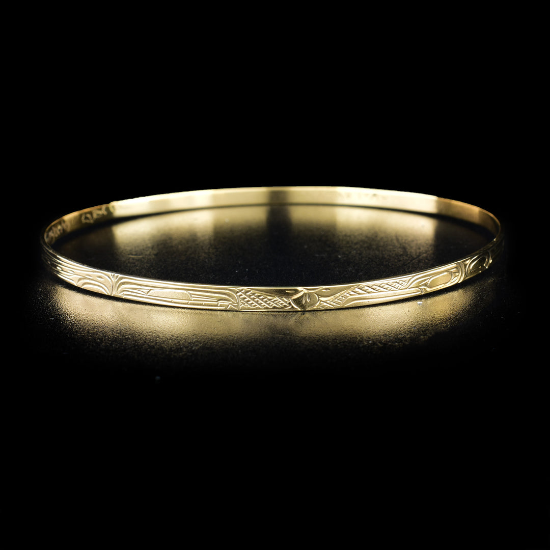 14K Gold 1/8" Orca, Hummingbird, and Wolf Bangle by Victoria Harper. The design depicts a hand carved flower in the center of the bracelet. To the right of the flower is the profile of a hummingbird's head facing towards the center. To the left of the flower there is a profile of an orca's head facing towards the center. The head of the wolf is located behind the hummingbird towards the back of the bangle.
