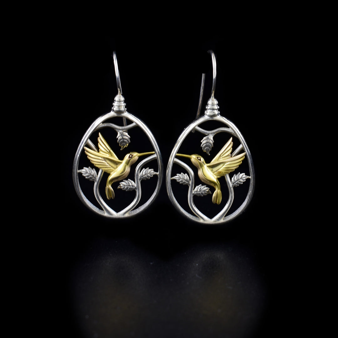 These garden hummingbird earrings are made from sterling silver and 14K gold. The hangs are oval shaped and have cutout and 3D designs depicting a hummingbird among leaves. The hummingbird is made from gold and the rest of the pice is silver.