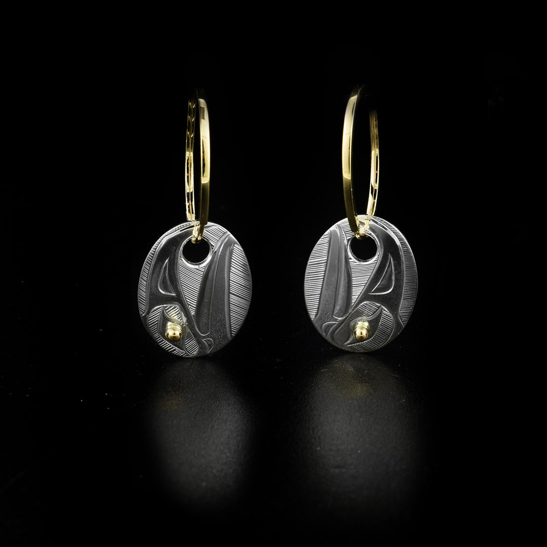 Gold and Silver Oval Raven Sleeper Hoop Earrings hand-carved by Haisla artist Hollie Bartlett. The dangling ravens are made of sterling silver and have 14K gold in their eyes. The hoops are made of 10K gold. Each earring measures 1.20" x 0.50" including sleeper hoops.