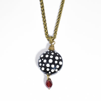Round, black handmade lampworked glass bead with white polka dots. Garnet hangs below. Metal is brass. Pendant is 2” x 0.81” including bail. Wheat chain is 17.5” long with 2.5” extension.