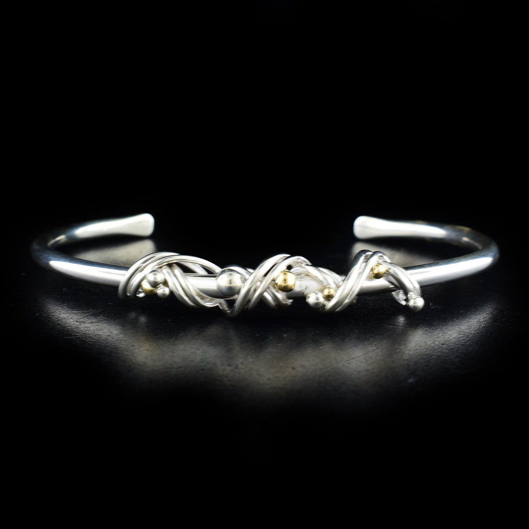 This silver and gold bangle bracelet has been made using sterling silver and 14K gold. The bangle is made from a silver rod with a gap in the back and has silver coil wrapped around the top of it. Featured on the coil are silver and gold beads.