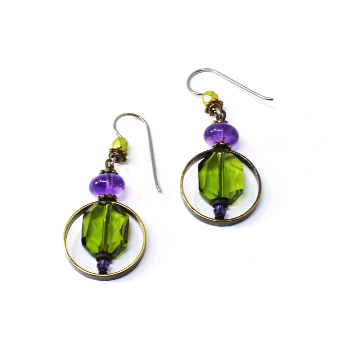These crystal drop earrings are made from Austrian crystal, amethyst, glass and antique brass. There is a small glass bead followed by two larger amethyst beads dangling from the hooks. Below the beads are large, green Austrian crystals with brass hoops wrapping around them.