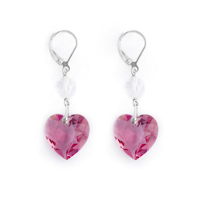 Pink Swarovski Crystal Heart Dangle Earrings by Debra Nelson. At the end of each earring is a pink Swarovski crystal in the shape of a heart. Above it is a clear Swarovski crystal in a round shape. The artist has included sterling silver accents that connect the crystals before attaching them on top sterling silver ear hooks.