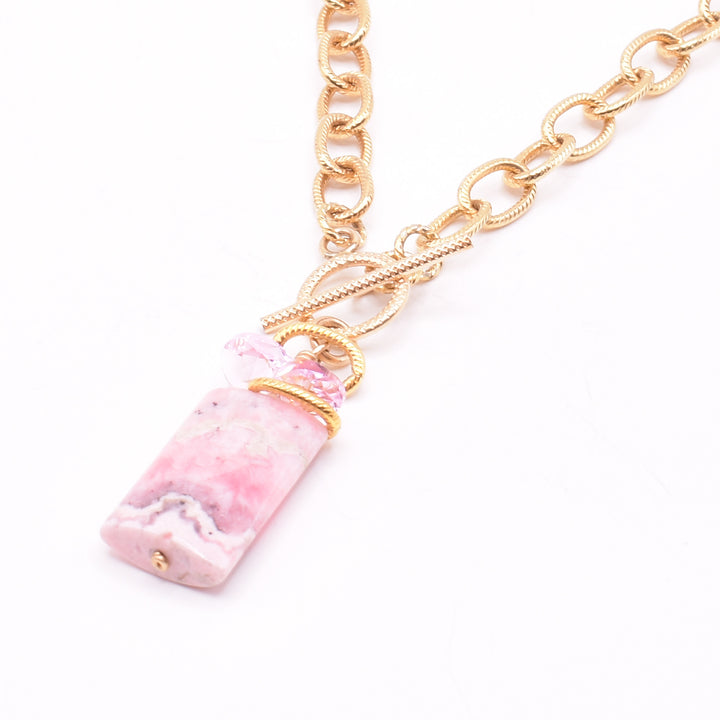 This crystal necklace with pendant is made rom 14K gold fill, pink Swarovski crystal and a large rhodonite gem. The pendant is wide, rectangular and made from rhodonite with swirling pink, designs on it. The chain is chunky and has circle shaped, gold links; there is pink cristal above the gemstone pendant.