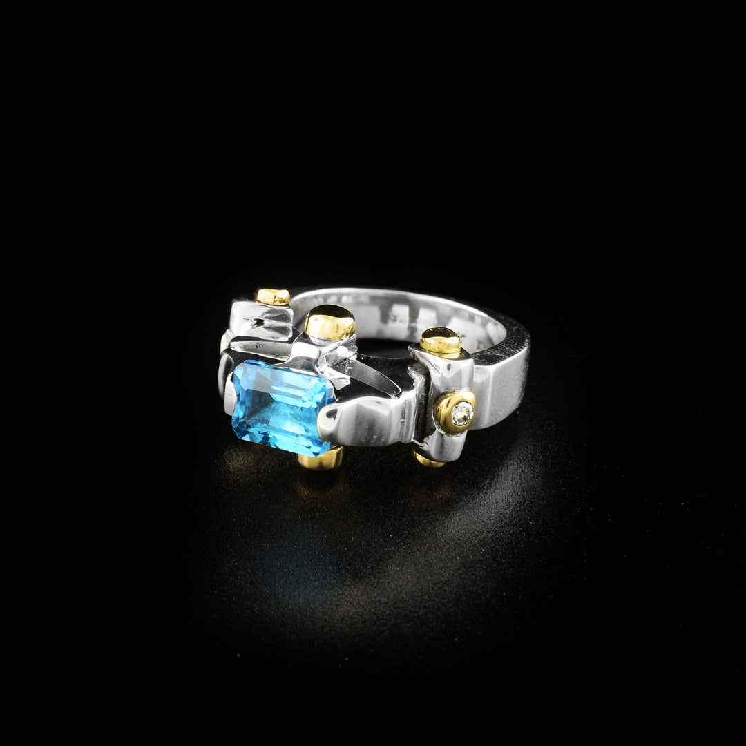 This blue topaz ring is made from sterling sliver, 14k gold, diamonds and a blue topaz gemstone. The silver band is thick and has a blue gemstone held with prongs. There are gold studs featured around the piece, and on either side of the blue gem are diamonds embedded into a stud.