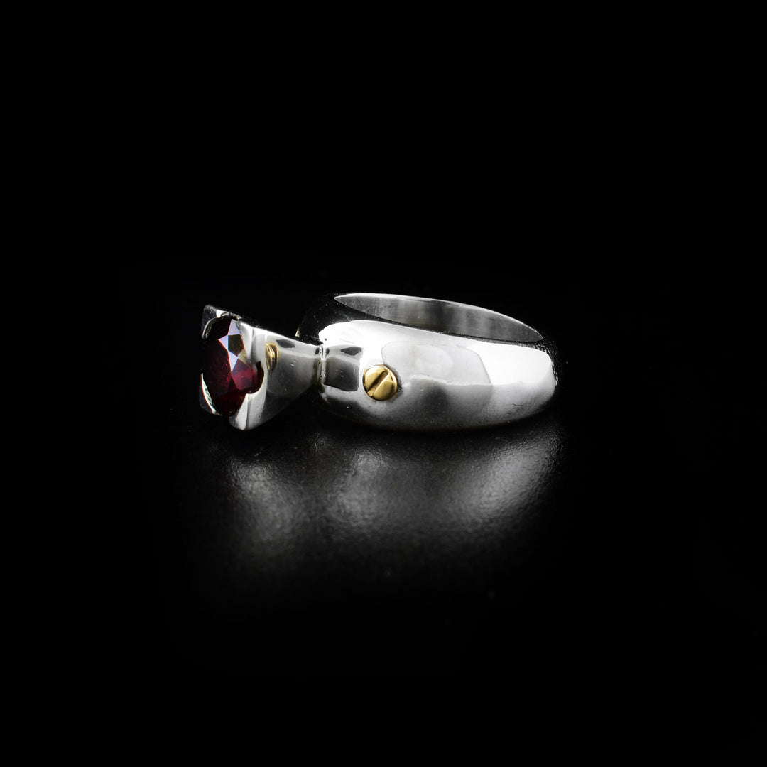 This garnet ring is made from sterling silver, 14K gold and garnet gemstone and has a single silver band. There is a garnet gemstone held in silver at the top of the band and gold studs/screws in the band on either side of the gem.