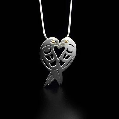 Silver and Gold Lovebirds Pendant by Grant Pauls. The pendant has two lovebirds facing towards each other, connecting to make a heart shape. Both lovebirds have a 18k gold accent in their eyes and intricate designs cut out along their bodies to represent their feathers.