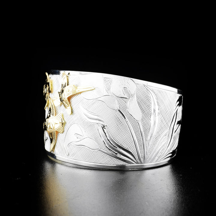 Hummingbird and calla lily bracelet hand-carved by Tlingit artist Fred Myra and contemporary Canadian artist Arlene Howk. 14K gold hummingbirds carved by Fred and sterling silver bracelet carved by Arlene. Bracelet is 6.75" long with 0.75" gap and is 1.50" wide. Ends are tapered.