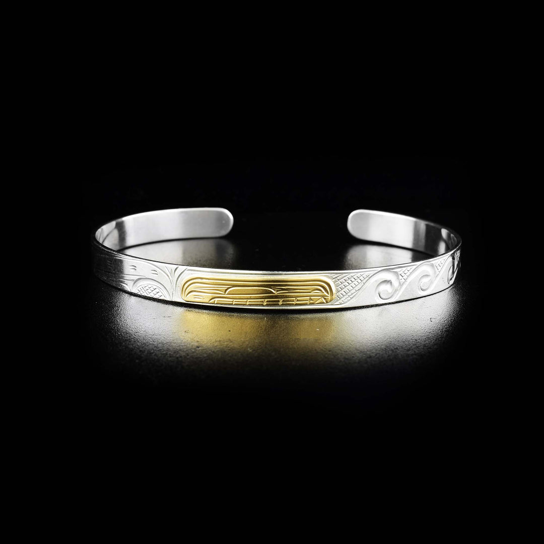 This silver and gold bangle bracelet is made from sterling silver, 14K gold and has a single, thin band with a gap in the back. There is a depiction of the Orca carved into the surface of the band and the head of the legend is made from gold.
