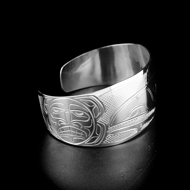 Tapered raven and sun bracelet hand-carved by Kwakwaka'wakw artist John Lancaster. Made of sterling silver. Bracelet is 6.15" long with 0.80" gap and has width of 1". Tapered ends.