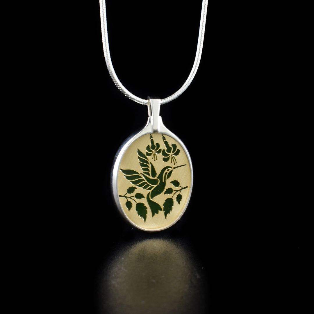 This brilliant hummingbird pendant is made from string silver 14K gold and grade A+ BC jade. There is a gold plate with handcarevd depictions of a hummingbird on it and it is backed with the jade. The piece is held in an oval band around the side of it and forming the bail.