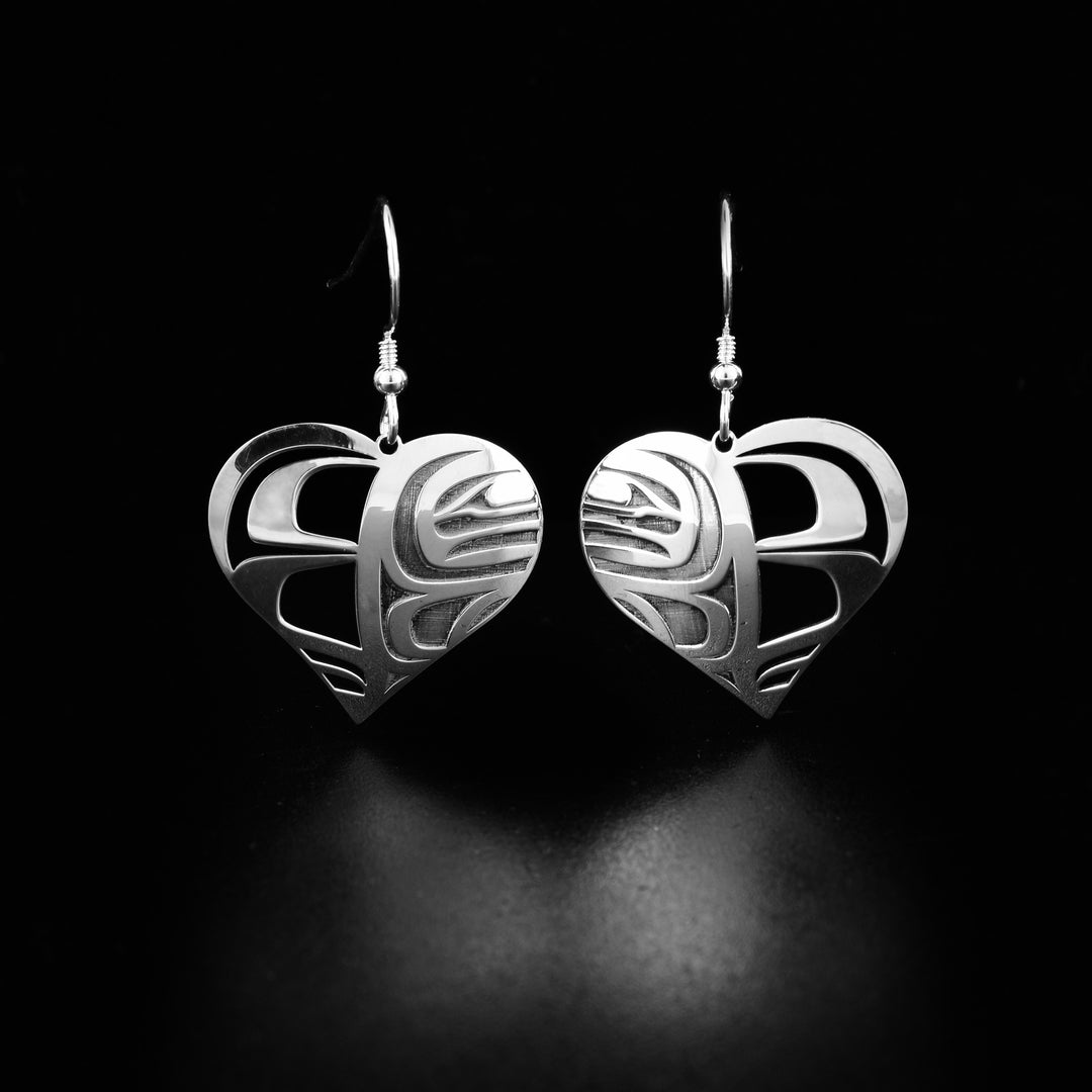 Orca cutout sterling silver heart earrings by Grant Pauls. Each earring is in the shape of a heart. Half of the heart has been cutout into intricate designs. The other half of the heart has been cut out to the face of an orca is visible.