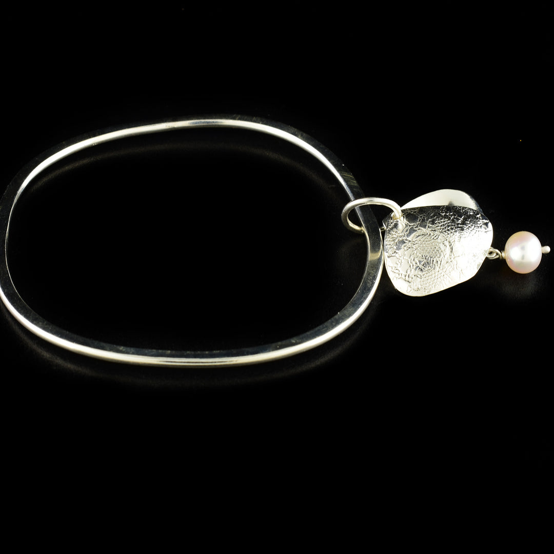 Unique pearl bangle handcrafted by artist Victoria Poynton. Made of sterling silver and a freshwater pearl. Victoria has used vintage French lace to emboss the design on the pendant. Bracelet has a circumference of 8" and pendant measures 1.50" x 0.88" including bail.