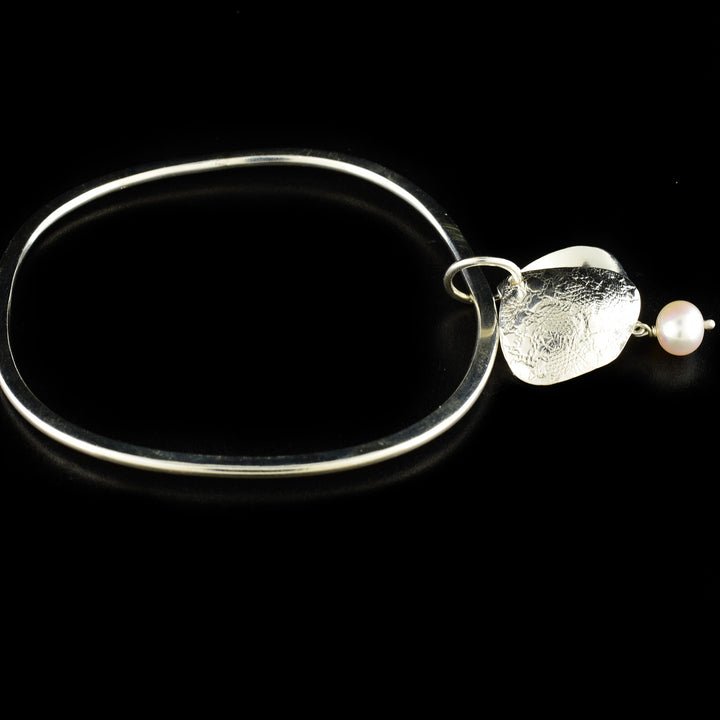 Unique pearl bangle handcrafted by artist Victoria Poynton. Made of sterling silver and a freshwater pearl. Victoria has used vintage French lace to emboss the design on the pendant. Bracelet has a circumference of 8" and pendant measures 1.50" x 0.88" including bail.
