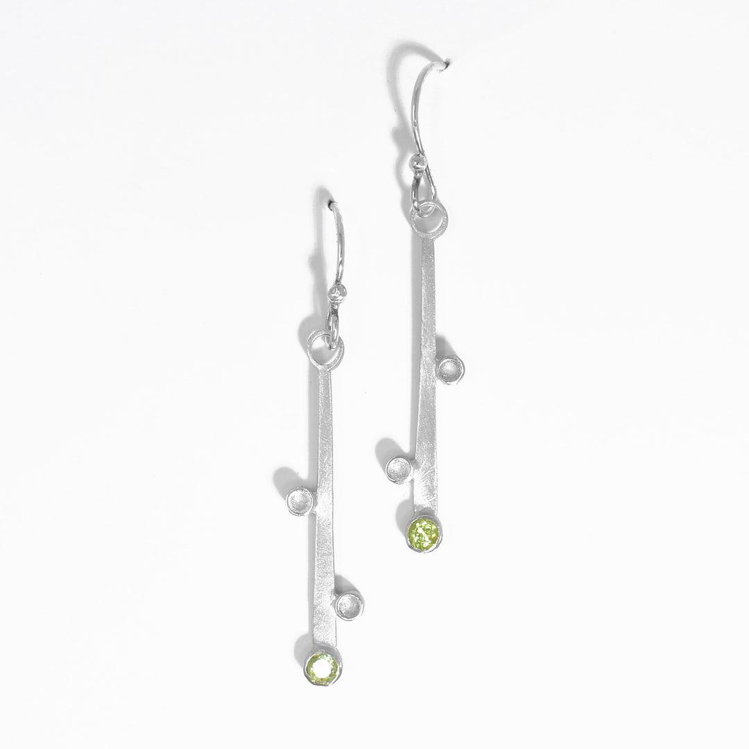Flat strips of silver with silver bobbles on sides hang from hooks. At end of each strip there is a peridot set in the metal. Each earring hangs 1.88” long including hook.