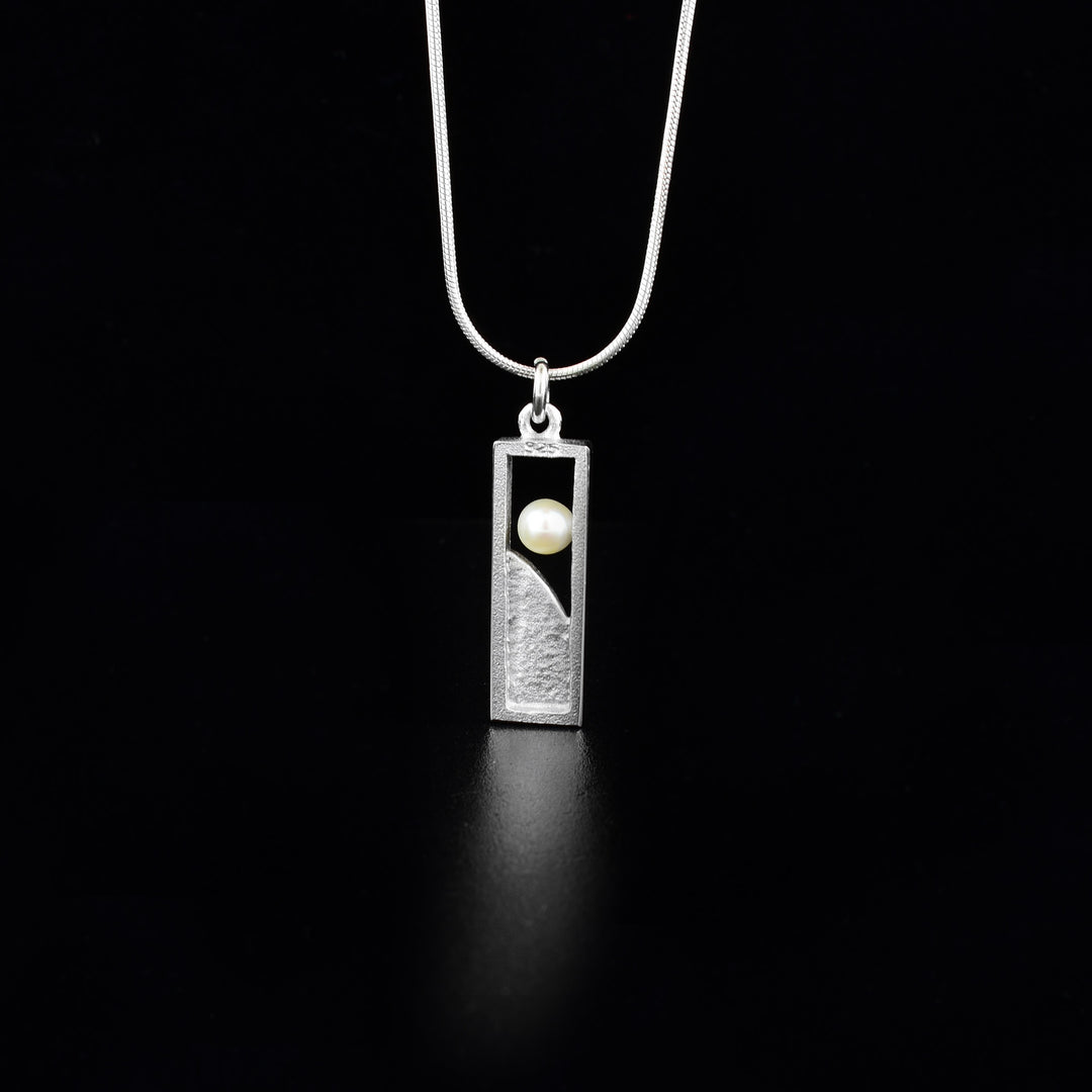 Sterling Silver Window Pearl Necklace by Jean Bastien. The pendant is in a rectangular shape. The bottom portion of the pendant has been filled in with sterling silver. The artist has included a white freshwater pearl towards the top of the necklace and has attached it to one of the sides of the pendant to give it a "floating effect". 