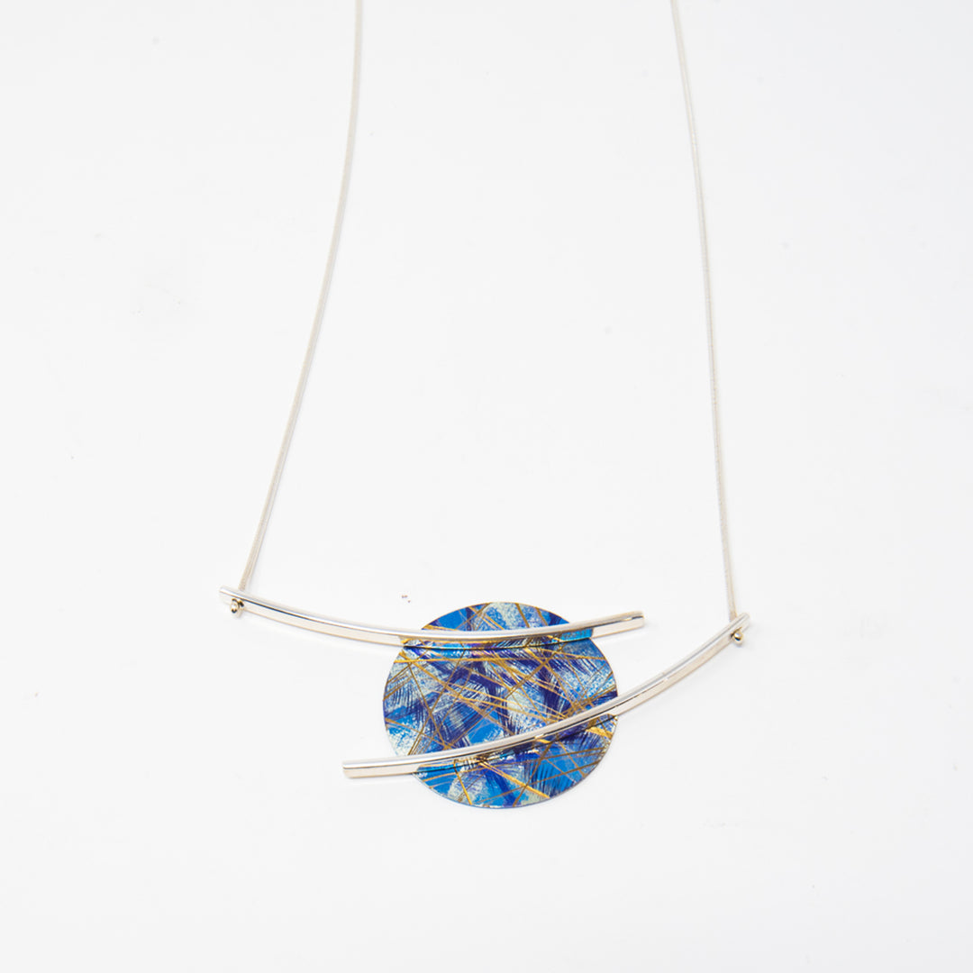 This titanium necklace is made from anodized titanium and sterling silver and has a circle shaped pendant. The circle is made from titanium and is blue with colorful designs that are achieved by anodizing the metal. There are sterling silver branches stretching across and past the pendant, giving it a floating appearance. 