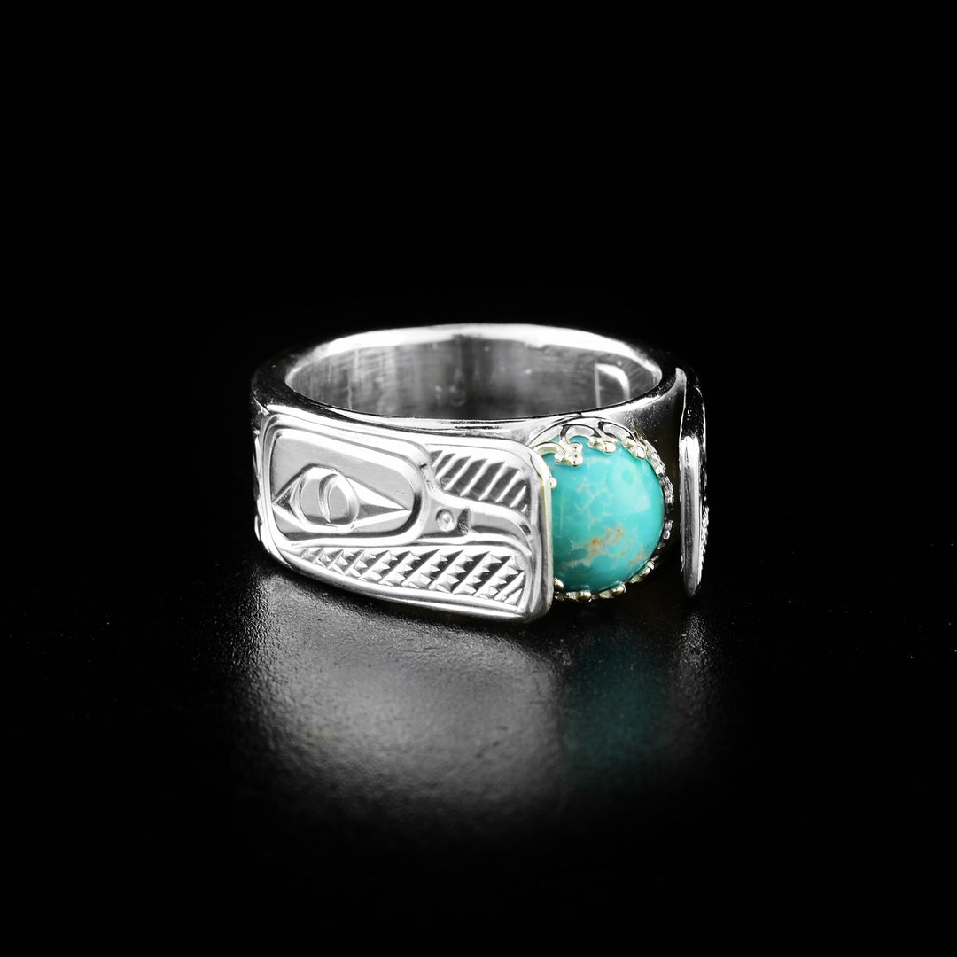 This wide cathedral ring has a wide, sterling silver band that extends up above the turquoise gem. The head of the Raven is carved into each side of the band and the beaks are facing towards the gem. Behind the head, there are feather designs carved. The turquoise gem is round and there are crown like designs that hold it into the piece.